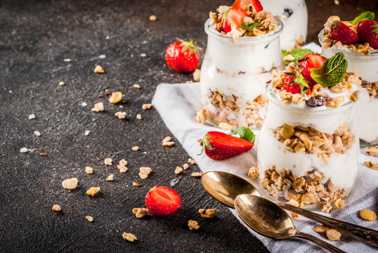 Healthy summer breakfast idea, homemade layered parfe dessert in small jar with granola, yoghurt and strawberry, dark background copy space