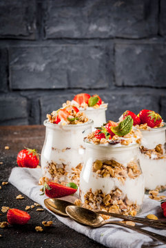 Healthy summer breakfast idea, homemade layered parfe dessert in small jar with granola, yoghurt and strawberry, dark background copy space