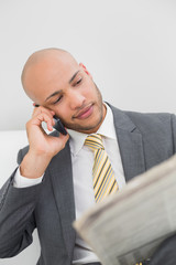 Elegant businessman using cellphone while reading newspaper