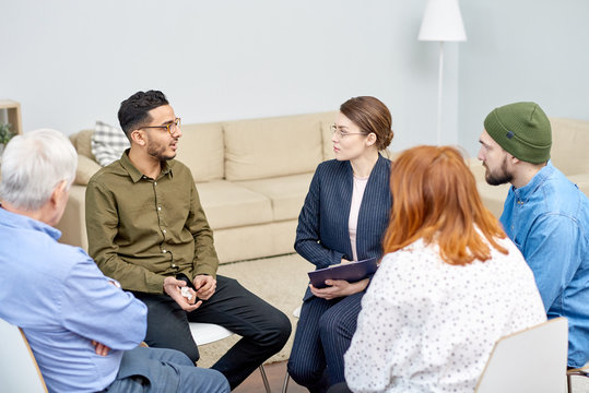 Depressed mixed-race patient with paper tissue in hand talking to highly professional psychologist while participating in group therapy session, interior of cozy office on background
