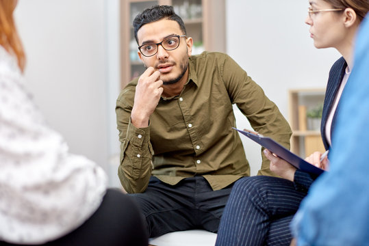 Portrait shot of handsome mixed-race man wearing eyeglasses participating in group therapy session at cozy office, friendly psychologist assisting her patients