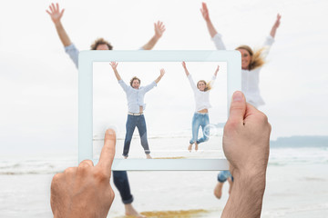 Hand holding tablet pc showing cheerful young couple jumping at beach