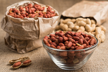 Glass bowl and paper bag full of peeled peanuts and peanuts in nutshell on sackcloth