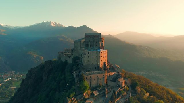 Aerial: drone flying at old medieval abbey perched on mountain top, background snowy Alps at sunrise. Sacra di San Michele (italian) - Saint Michel Abbey (english traslation) - Turin, Italy