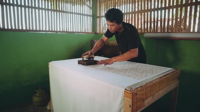 Adult indonesian man applying hot wax pattern to fabric using cap to make batik