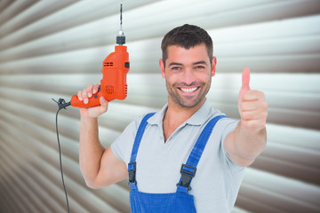 Smiling repairman with drill machine gesturing thumbs up against grey shutters