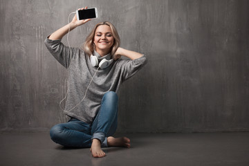 Young and attractive woman with smartphone and large headphones, sitting on the floor. Music in the mobile app.