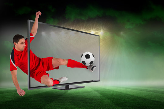 Composite image of fit football player kicking ball through tv against football pitch under green sky and spotlights