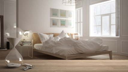 Wooden table, desk or shelf with crystal modern hourglass measuring the passing time in a countdown to a deadline and house keys over modern bedroom, architecture interior design