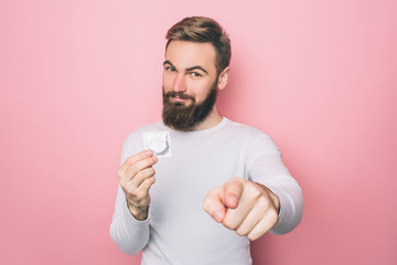 Happy man is holding a condom and pointing on camera. He is smiling. Isolated on pink background.