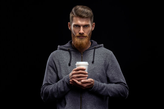 Dramatic light studio shot of young caucasian bearded stylish man with cup of coffee Handsome hairstyling male portrait holding coffee to go cup over black background .