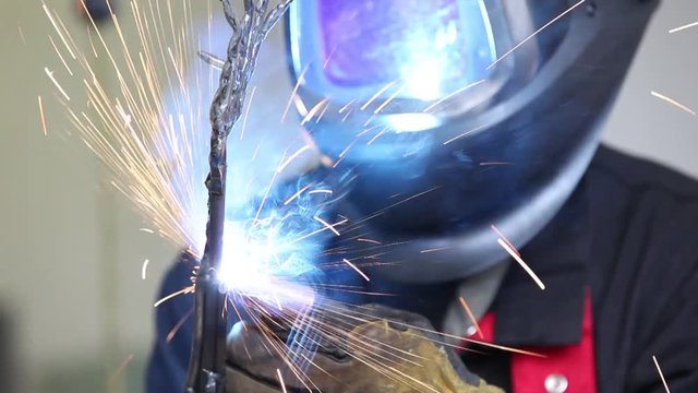 A front view of a man wearing a welding helmet and gloves using an arc welder on a metal armature, producing sparks and light which is reflected in man's visor.