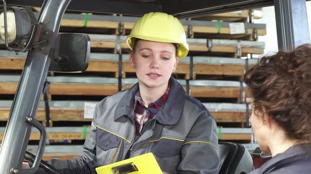Attractive female forklift operator working at the storage wearing protective hardhat talking to her colleague. Professionalism, partnership, distribution, emancipation concept.