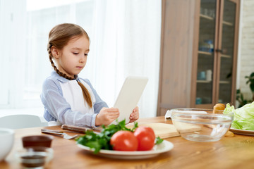 Adorable little girl wearing apron browsing Internet on digital tablet in order to find recipe of appetizing starter while wrapped up in making surprise for birthday of her dad