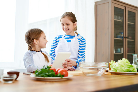 Cheerful little sisters wearing aprons looking at each other with wide smiles while preparing appetizing starter for their parents, spacious kitchen interior on background