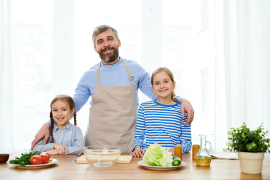 Handsome bearded man wearing apron embracing his little daughters while looking at camera with toothy smile, interior of modern kitchen on background