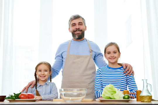 Cheerful bearded man and his little daughters looking at camera with wide smiles while gathered together at spacious kitchen and making delicious surprise for mom, group portrait shot