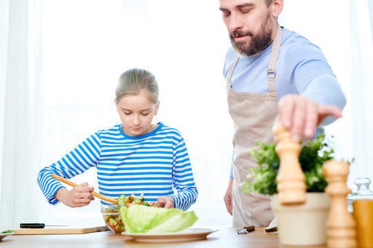 Making pleasant surprise for mom: pretty little girl preparing delicious salad while her bearded dad assisting her, kitchen interior on background