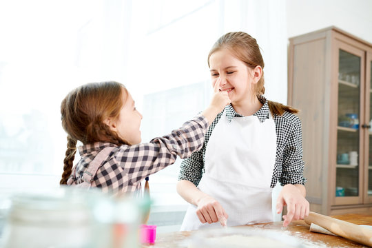 Profile view of cute little girl smearing face of her elder sister with flour while preparing pie together, interior of spacious kitchen on background