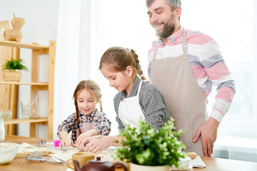 Pretty little sisters cutting cookies from dough while standing at wooden kitchen table, their smiling bearded dad keeping eye on them