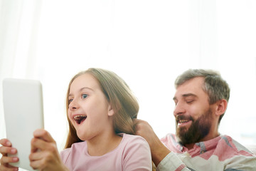 Funny little girl taking selfie with bearded dad on digital tablet while he combing her hair, close-up shot