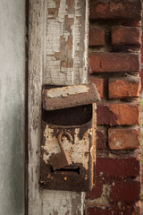 Rusty mailbox mounted on a wood door frame with peeling, cracked, white paint, next to an old brick wall