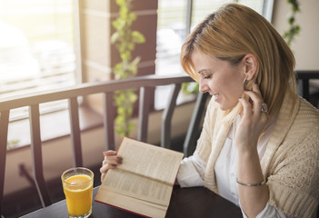 Young smiling blonde woman in a restaurant reading a book and drinking juice