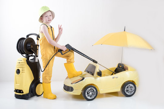 Happy child in yellow overalls and boots with high pressure washer serves client boy on toy car under umbrella. Concept of family business. White background