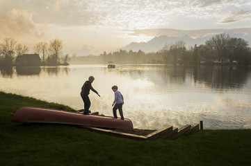 Two young boys play on canoe on beach of lake at sunset
