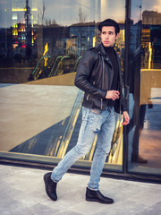Obraz premium One handsome young man in urban setting in modern city, walking, wearing black leather jacket and jeans