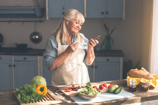 Relaxed mature woman is choosing song on smartphone playlist and smiling. She is standing in kitchen and wearing earphones during lunch preparation