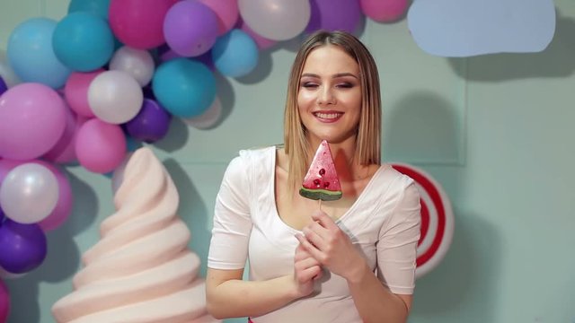 Portrait of a happy girl with a big candy in the form of watermelon on a background of multi-colored balls and giant ice cream. Barbie. Lollipop.
