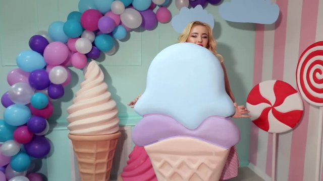 A cheerful cute girl holds a huge ice cream in the studio on a background of giant sweets. Barbie style. The girl is a sweet tooth.