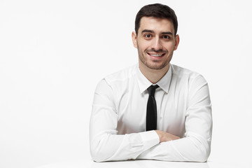 Indoor picture of handsome Caucasian male dressed in white shirt and necktie pictured isolated on white background resting arms on white table feeling optimistic and ready for business communication