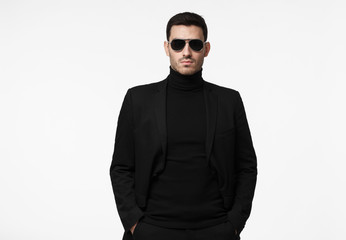 Obraz premium Half-length portrait of young handsome European guy isolated on white background dressed in total black with turtleneck, sunglasses and jacket, looking serious and suspicious, reserved and reticent