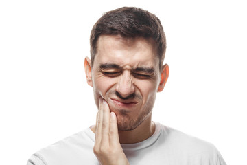 Indoor portrait of young handsome Caucasian guy pictured isolated on white background, his face distorted and hand pressed to cheek as he is experiencing severe toothache and worries about it