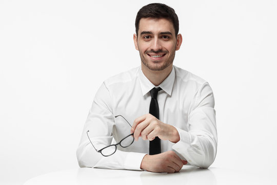 Closeup photo of handsome Caucasian male pictured isolated on white background in white shirt and necktie resting arms on desk, holding glasses in hand, listening to partner with interest and content