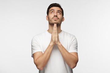 Indoor picture of young European Caucasian man pictured isolated on gray background dressed in white T-shirt praying with hands pressed together, looking upwards as if asking god for blessing