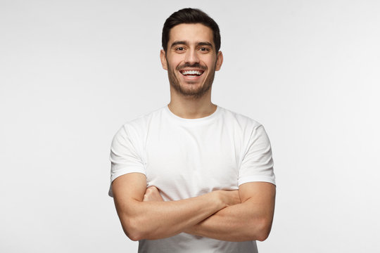 Half-length indoor portrait of young European Caucasian man isolated on gray background standing in white T-shirt with arms crossed, smiling and laughing positive as he is looking straight at camera