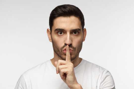 Horizontal photo of handsome European man isolated on gray background dressed in white casual T-shirt pressing finger to lips as if asking to keep silence, not willing anyone to know secret
