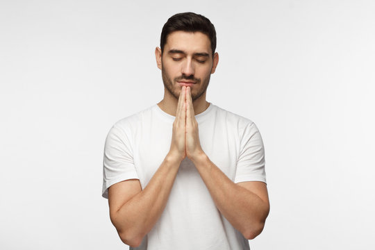 Closeup picture of handsome European man isolated on grey background in white casual T-shirt standing with closed eyes and hands pressed together as if meditating or praying, looking peaceful
