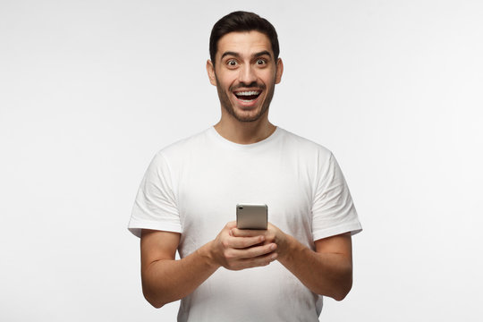 Horizontal photo of young good-looking European man isolated on grey background standing in white T-shirt holding cellphone in hands, eyes round with surprise, laughing joyfully as if having won prize