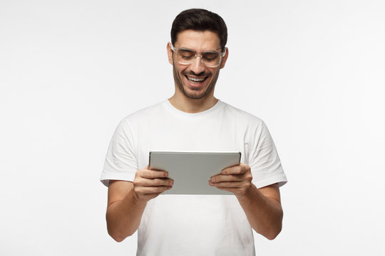Closeup picture of good-looking European man pictured isolated on grey background looking through trendy transparent glasses at screen of tablet computer he is holding in hands in front of him