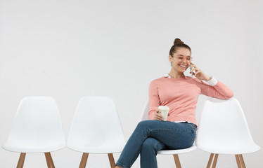 Indoor shot of smiling female student sitting in the line with coffee cup, glad to hear boyfriend over smart phone