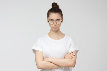Fototapeta premium Closeup picture of pretty European Caucasian girl pictured isolated on grey background with white T-shirt and glasses, looking seriously at camera with arms crossed as if suspicious showing mistrust