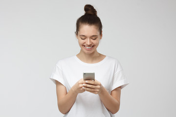Fototapeta premium Horizontal portrait of young positive European Caucasian woman isolated on grey background wearing white T-shirt, looking with smile at screen of smartphone, exchanging messages or browsing web pages