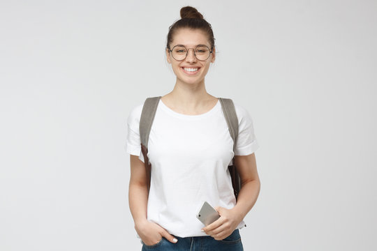 Horizontal photo of young beautiful European woman pictured isolated on grey background wearing round glasses and bagpack, smiling happily, holding smartphone in one hand, ready to start journey