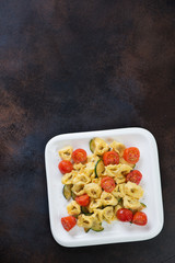 White plate with italian tortellini, fried zucchini and tomatoes. Above view on a dark brown metal surface, vertical shot with copyspace