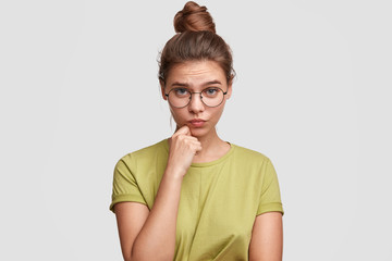 Horizontal portrait of lovely blue eyed female student with hair knot, wears big round spectacles, holds chin, looks seriously and with shy expression at camera, isolated on white background.