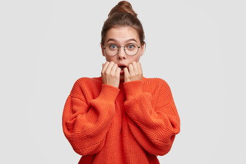 Pretty nervous young female has worried expression, feels anxious, looks with blue eyes surprisingly, wears round big spectacles and oversized red knitted sweater, isolated over white background